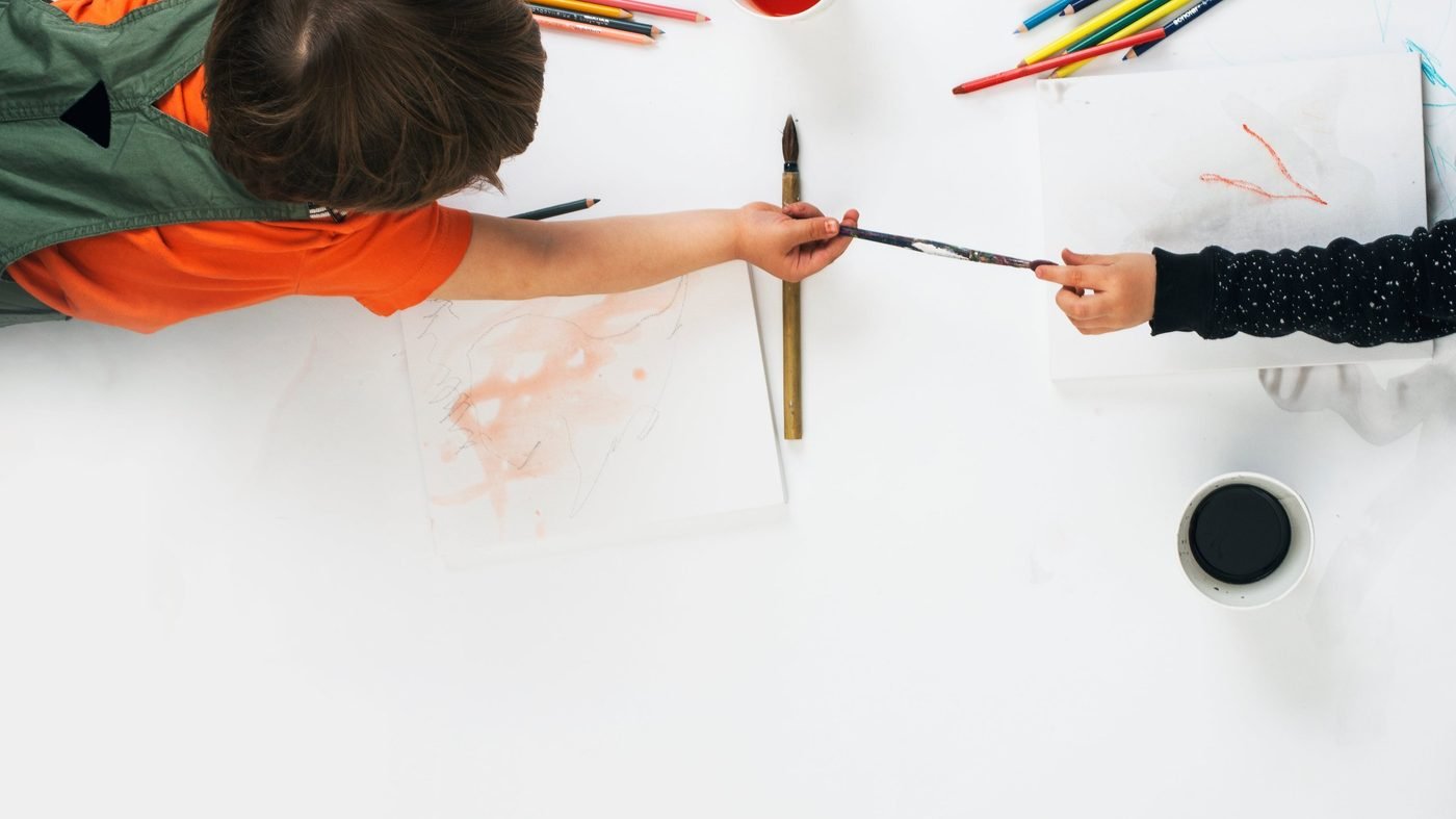 Two children at a white table covered in art supplies, one passing a paintbrush to the other